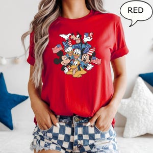 May include: Red t-shirt featuring a graphic of Disney characters, including Mickey Mouse, Minnie Mouse, Donald Duck, Goofy, and Pluto, holding American flags. The word "RED" is in a speech bubble. The shirt is red.