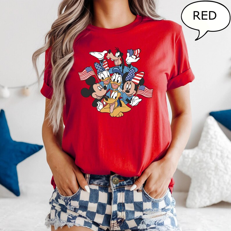 May include: Red t-shirt featuring a graphic of Disney characters, including Mickey Mouse, Minnie Mouse, Donald Duck, Goofy, and Pluto, holding American flags. The word "RED" is in a speech bubble. The shirt is red.