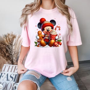 Puede incluir: Camiseta rosa con un gr&aacute;fico navide&ntilde;o de Mickey Mouse con un gorro y una bufanda rojos, sosteniendo una taza de chocolate caliente y un hombre de jengibre.