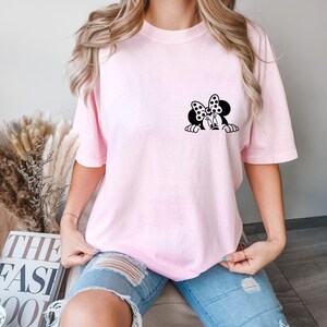 May include: A pink t-shirt with a black and white graphic of Minnie Mouse peeking over the top. Minnie Mouse is wearing a bow with polka dots.