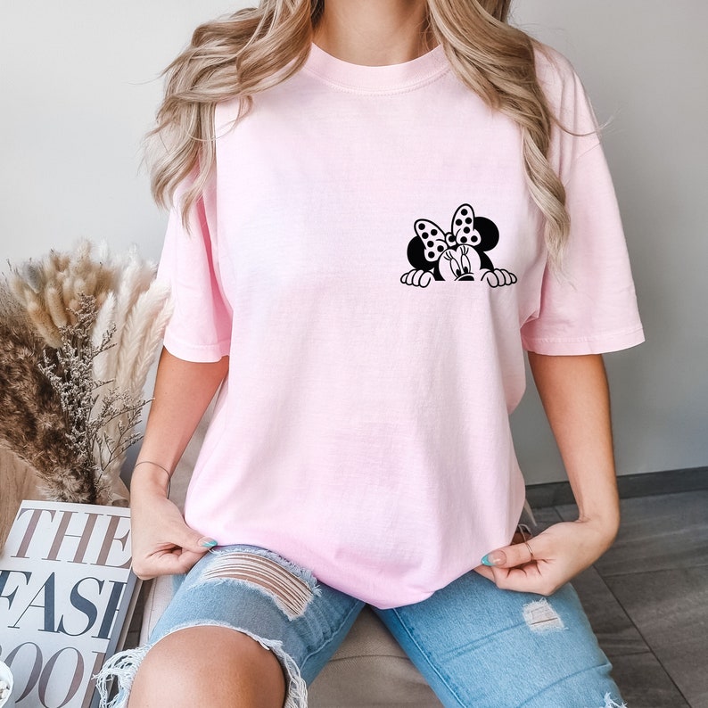 May include: A pink t-shirt with a black and white graphic of Minnie Mouse peeking over the top. Minnie Mouse is wearing a bow with polka dots.