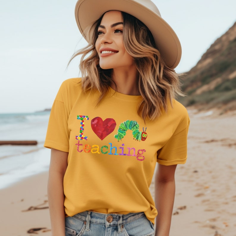 Op de afbeelding: Een vrouw draagt een geel T-shirt met de tekst "Ik hou van lesgeven" en een kleurrijke rupsafbeelding. Het shirt is lichtgeel en de tekst is in regenboogkleuren. De vrouw draagt een strohoed en staat op een strand.
