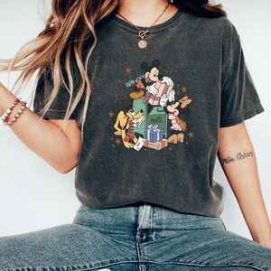 May include: A dark grey t-shirt featuring a vintage-style graphic of Mickey Mouse, Minnie Mouse, and Pluto delivering mail and gifts. The graphic includes a green mailbox with the word "Mail" on it, and the characters are holding parcels and presents.