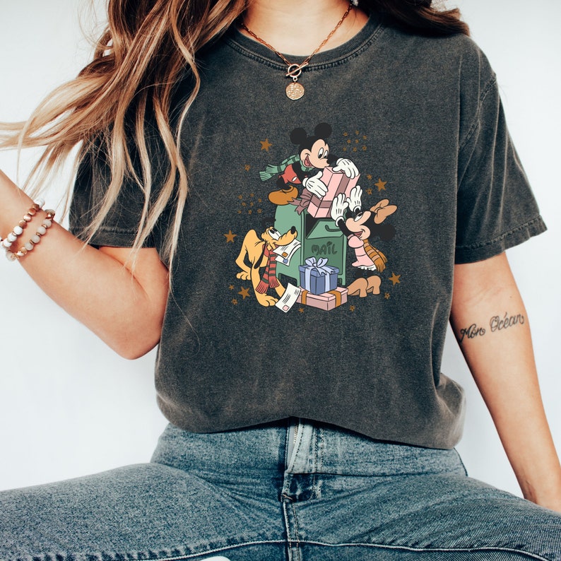 May include: A dark grey t-shirt featuring a vintage-style graphic of Mickey Mouse, Minnie Mouse, and Pluto delivering mail and gifts. The graphic includes a green mailbox with the word "Mail" on it, and the characters are holding parcels and presents.