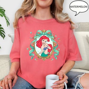 May include: Coral-colored t-shirt featuring a framed illustration of a red-haired mermaid with a blue seashell top and green tail. The frame is gold with teal ribbons and floral accents. The word "WATERMELON" is in a speech bubble.