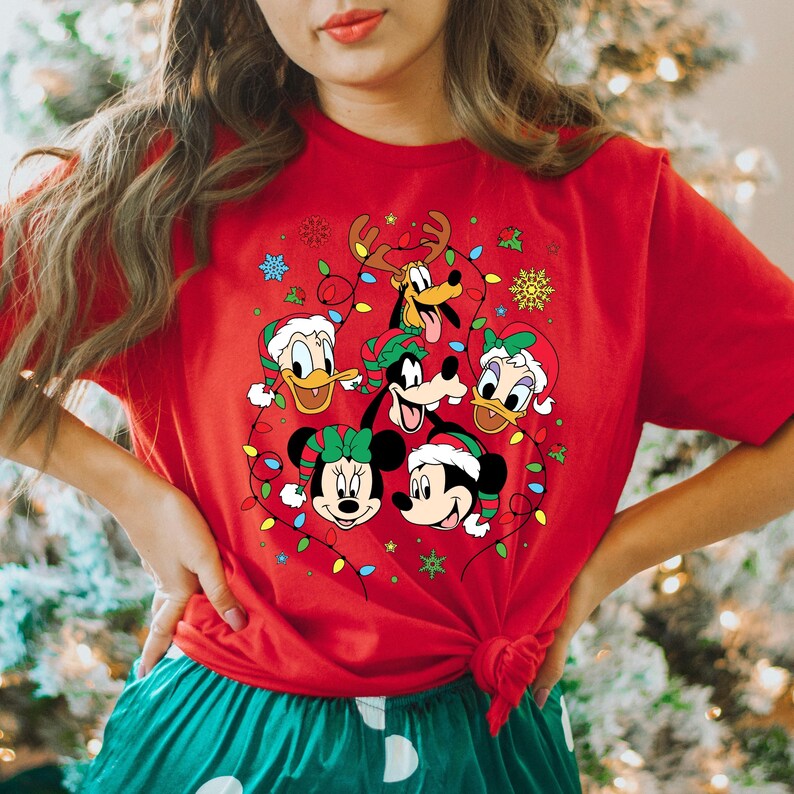 May include: Red t-shirt with a Christmas-themed design featuring Mickey Mouse, Minnie Mouse, Donald Duck, Goofy, and Pluto. The characters are surrounded by Christmas lights and snowflakes.