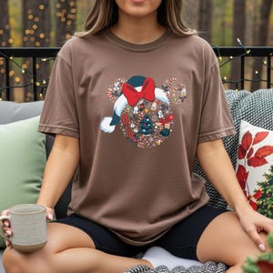 Puede incluir: Camiseta marr&oacute;n con un dise&ntilde;o festivo. El gr&aacute;fico incluye un gorro de Pap&aacute; Noel con un lazo rojo y una disposici&oacute;n circular de elementos navide&ntilde;os. El dise&ntilde;o incorpora personajes de dibujos animados, un &aacute;rbol de Navidad y copos de nieve. La camiseta es de corte c&oacute;modo.