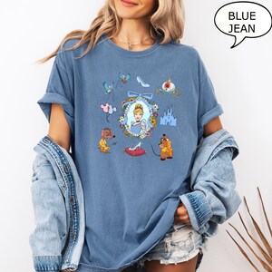May include: A blue jean t-shirt with a graphic print of Cinderella, her mice friends, and other elements from the Disney movie Cinderella. The graphic includes a glass slipper, a castle, and a carriage.
