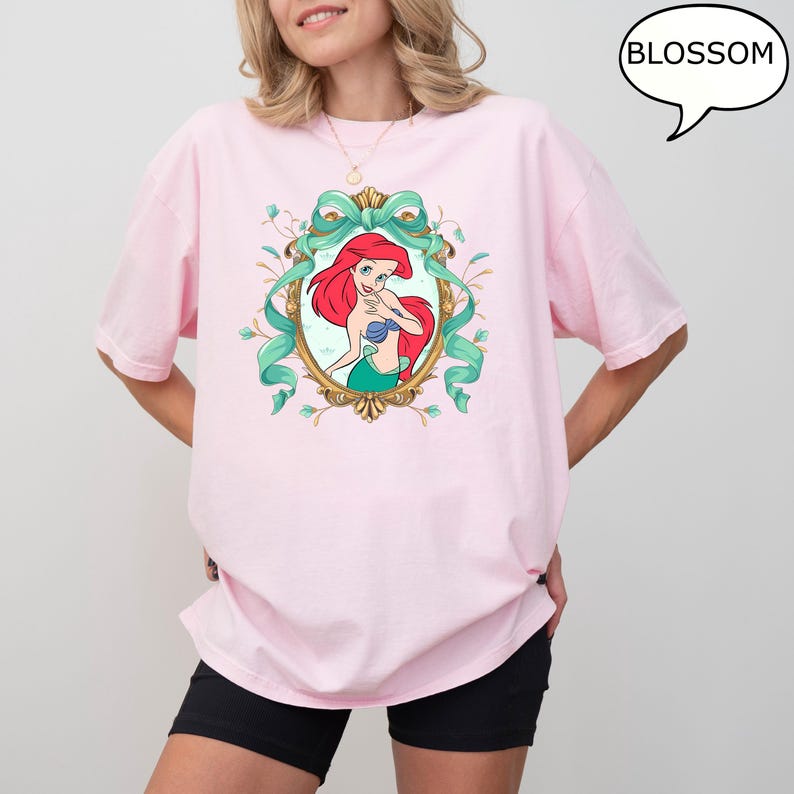 May include: A light pink t-shirt featuring a framed illustration of Ariel from The Little Mermaid. The illustration is surrounded by a teal ribbon and floral accents. The word "BLOSSOM" is in a speech bubble.