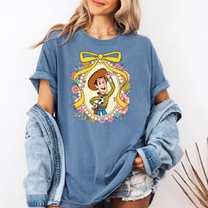 May include: A blue t-shirt with a cartoon image of Woody from Toy Story. Woody is wearing his signature cowboy hat and vest. He is surrounded by a floral wreath with a yellow bow.