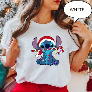 Puede incluir: Camiseta blanca con un personaje de dibujos animados, Stitch, con un gorro de Pap&aacute; Noel y sosteniendo un bast&oacute;n de caramelo. El personaje es azul con orejas grandes y luces navide&ntilde;as. La palabra "WHITE" est&aacute; en una burbuja.