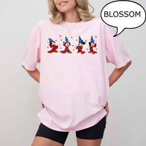 May include: A light pink t-shirt featuring a cartoon design of Mickey Mouse dressed as a wizard. The design includes four Mickey Mouse figures in red robes and blue wizard hats, with stars. The word "BLOSSOM" is in a speech bubble.