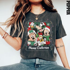 May include: A dark gray t-shirt with a vintage-style photo of Mickey Mouse, Minnie Mouse, Goofy, and Donald Duck wearing Santa hats. The photo is surrounded by a string of Christmas lights and the text "Merry Christmas".