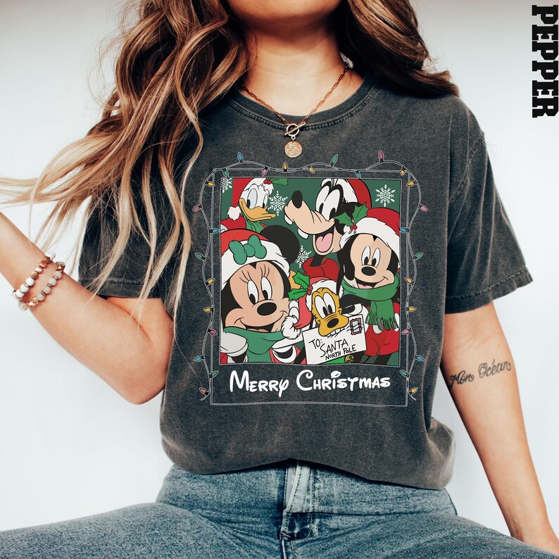 May include: A dark gray t-shirt with a vintage-style photo of Mickey Mouse, Minnie Mouse, Goofy, and Donald Duck wearing Santa hats. The photo is surrounded by a string of Christmas lights and the text "Merry Christmas".
