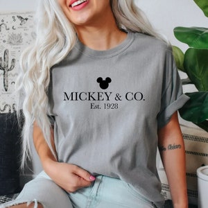Puede incluir: Camiseta gris con una cabeza de Mickey Mouse negra y el texto "MICKEY & CO. Est. 1928"