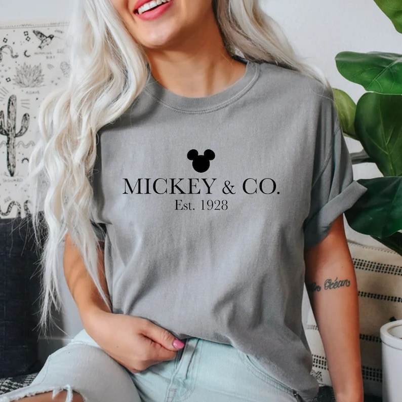Puede incluir: Camiseta gris con una cabeza de Mickey Mouse negra y el texto "MICKEY & CO. Est. 1928"
