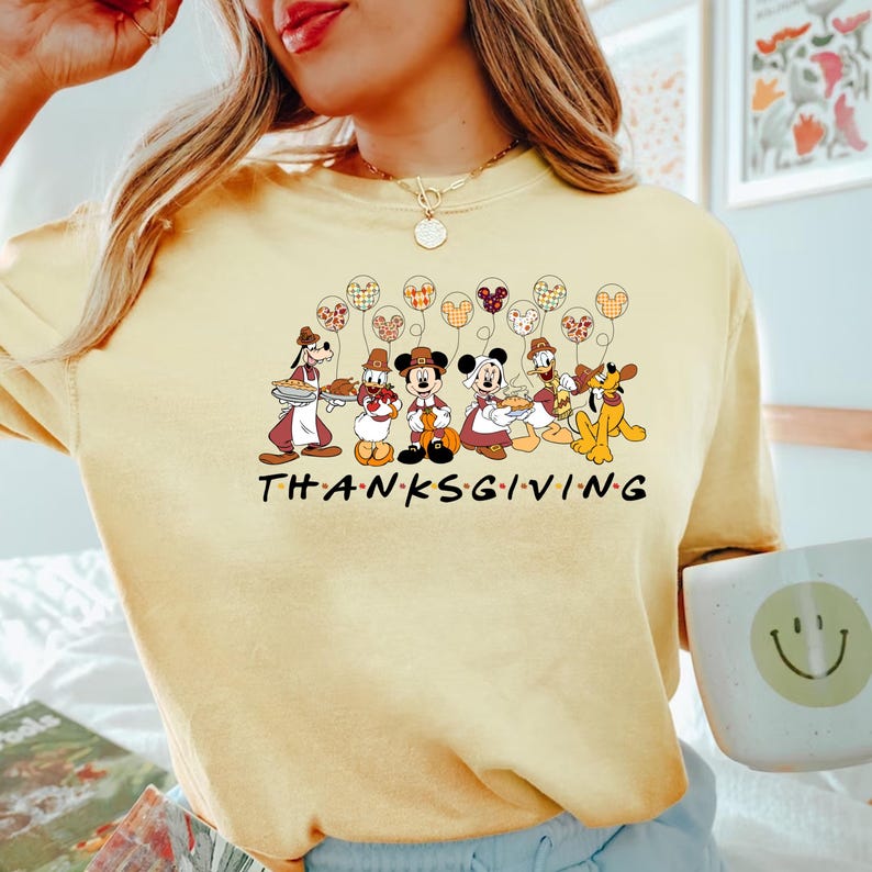 May include: A yellow t-shirt with a Thanksgiving theme featuring Mickey Mouse and friends. The shirt has a graphic of Mickey Mouse, Minnie Mouse, Goofy, Pluto, and Donald Duck dressed in Thanksgiving attire. The text "THANKSGIVING" is printed below the graphic.