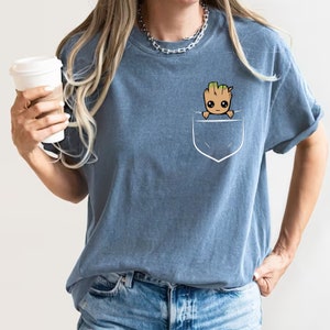 May include: A blue t-shirt with a pocket featuring a cartoon image of a small, brown, tree-like creature with green leaves and a smiling face. The creature is peeking out of the pocket.
