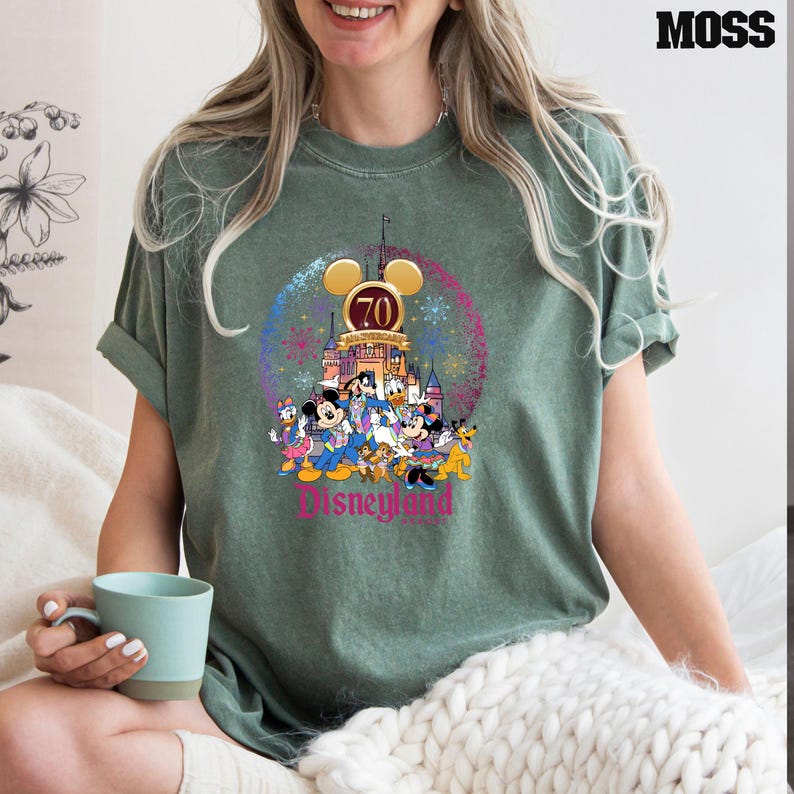 May include: A green t-shirt with a vintage-style graphic of Mickey Mouse and friends celebrating Disneyland's 70th anniversary. The graphic features a castle, fireworks, and the text "Disneyland 70th Anniversary".