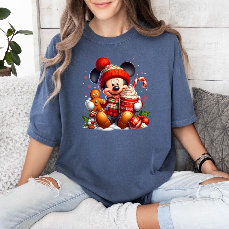 Puede incluir: Una camiseta azul con una imagen navide&ntilde;a de Mickey Mouse sosteniendo una taza de chocolate caliente y un hombre de jengibre. Mickey lleva un gorro y una bufanda de Pap&aacute; Noel rojos y blancos.