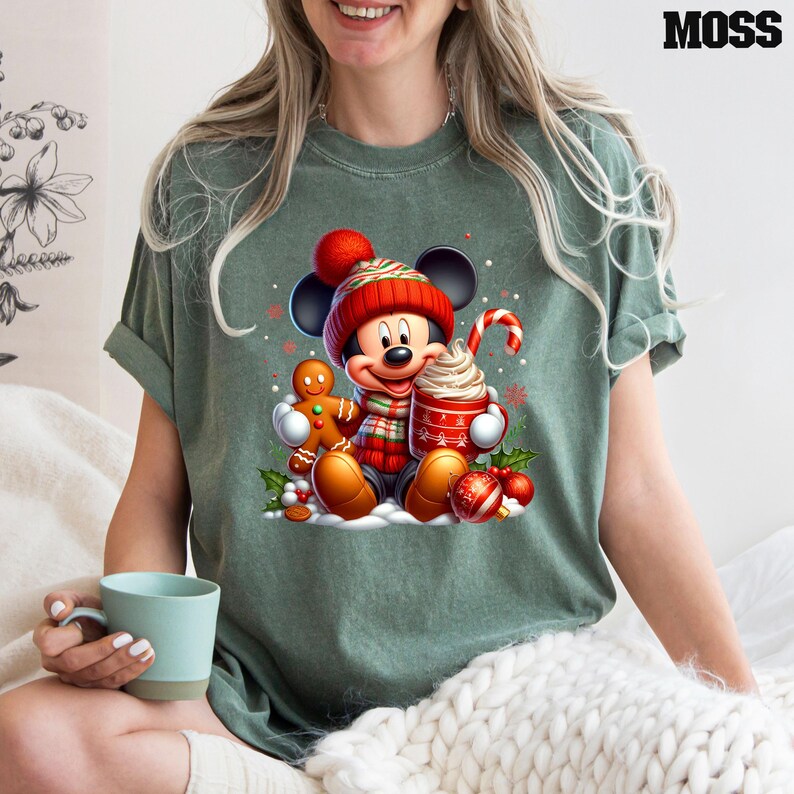 Puede incluir: Una camiseta verde con un gr&aacute;fico navide&ntilde;o que presenta a Mickey Mouse con un gorro de Pap&aacute; Noel rojo, sosteniendo una taza de chocolate caliente con crema batida y un bast&oacute;n de caramelo. Mickey est&aacute; de pie junto a un hombre de jengibre y rodeado de decoraciones navide&ntilde;as.