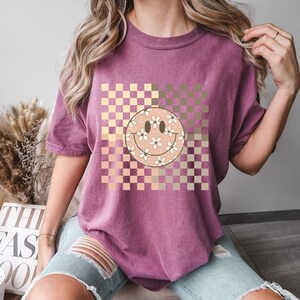 Peut inclure: Un t-shirt violet avec un motif &agrave; carreaux dans des tons de rose et de brun. Un grand visage souriant avec des fleurs blanches est centr&eacute; sur le motif &agrave; carreaux.