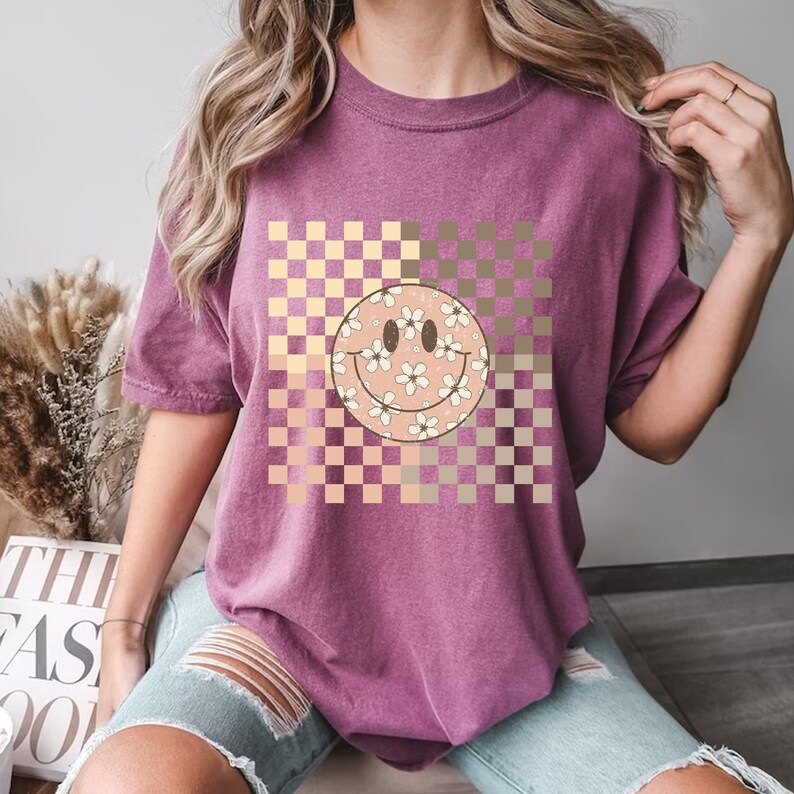 Peut inclure: Un t-shirt violet avec un motif &agrave; carreaux dans des tons de rose et de brun. Un grand visage souriant avec des fleurs blanches est centr&eacute; sur le motif &agrave; carreaux.