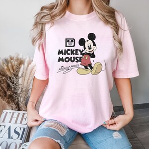 May include: A pink t-shirt with a vintage Mickey Mouse graphic. The graphic features Mickey Mouse in his signature red shorts and yellow shoes. The text "Mickey Mouse" is printed in black below the graphic.