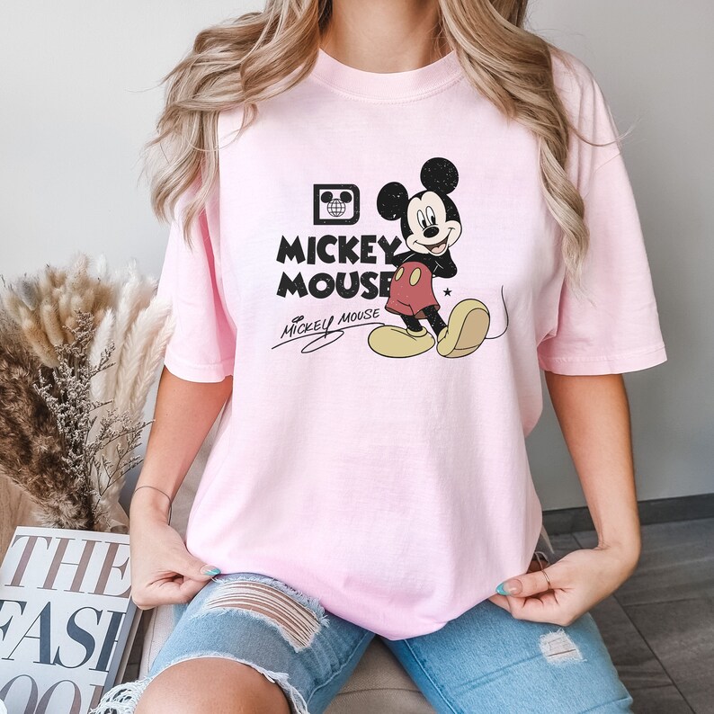May include: A pink t-shirt with a vintage Mickey Mouse graphic. The graphic features Mickey Mouse in his signature red shorts and yellow shoes. The text "Mickey Mouse" is printed in black below the graphic.