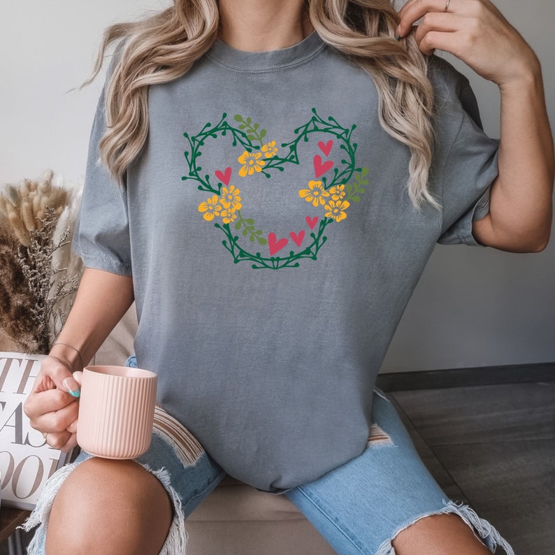 K&ouml;nnte beinhalten: Ein graues T-Shirt mit einem floralen Herzdesign. Das Herz besteht aus gr&uuml;nen Ranken mit gelben Blumen und roten Herzen.