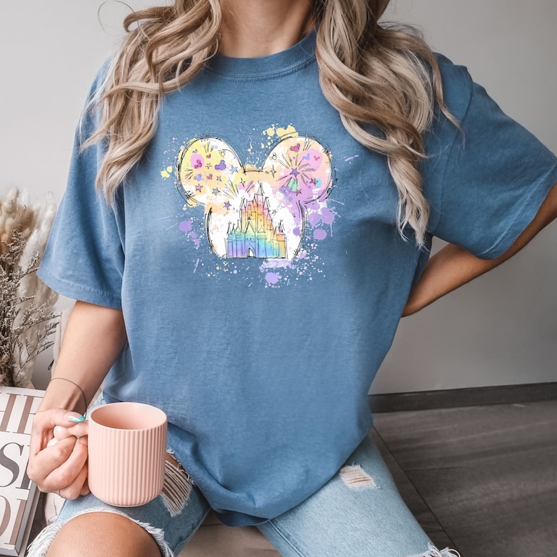 Puede incluir: Una camiseta azul con un gr&aacute;fico colorido de un castillo dentro de una silueta de cabeza de Mickey Mouse. El gr&aacute;fico tiene un efecto acuarela con salpicaduras de rosa, amarillo y azul.