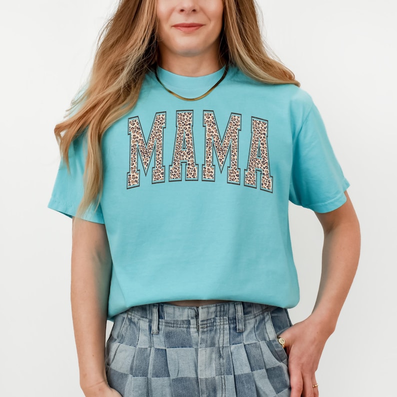 May include: A turquoise blue t-shirt with a large "MAMA" graphic in a leopard print design. The letters are outlined in black.
