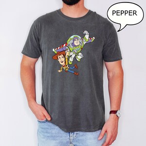 May include: Dark grey t-shirt featuring a colorful graphic of Buzz Lightyear and Woody from Toy Story. The characters are depicted in mid-air, with Buzz Lightyear in his signature space suit and Woody in his cowboy attire. The word "PEPPER" is in a speech bubble.