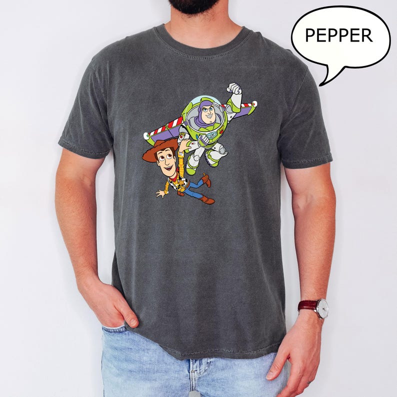 May include: Dark grey t-shirt featuring a colorful graphic of Buzz Lightyear and Woody from Toy Story. The characters are depicted in mid-air, with Buzz Lightyear in his signature space suit and Woody in his cowboy attire. The word "PEPPER" is in a speech bubble.