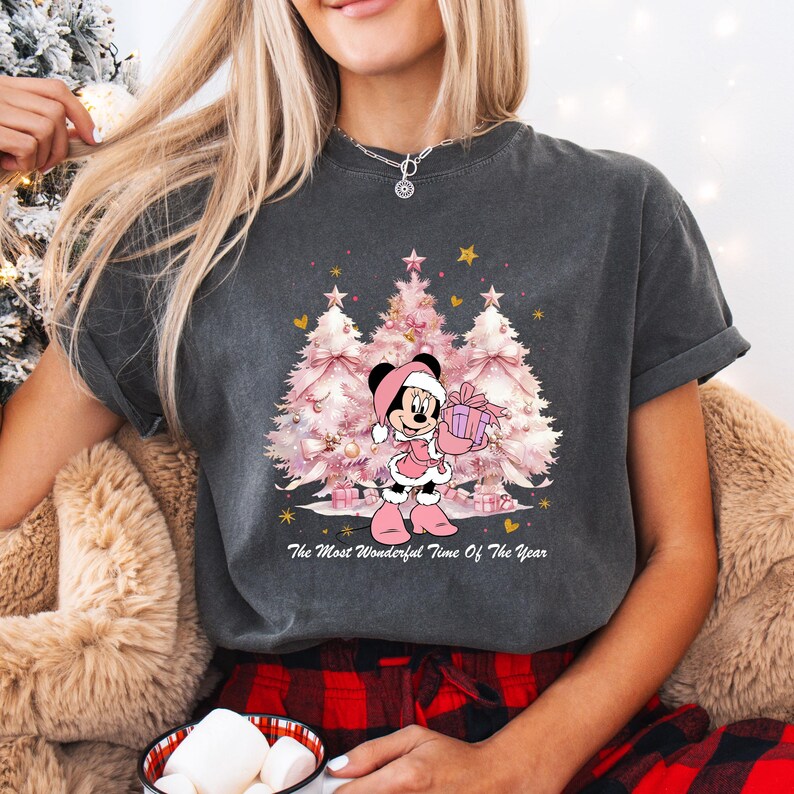 May include: Dark gray t-shirt featuring a Christmas design. The graphic includes pink Christmas trees, gold stars, and a cartoon character in a Santa hat holding a gift. Text reads "The Most Wonderful Time Of The Year."