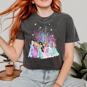 Puede incluir: Una camiseta gris oscuro recortada con un estampado gr&aacute;fico del castillo de Disney y siete princesas Disney. Las princesas llevan vestidos coloridos y est&aacute;n de pie frente al castillo. Hay corazones rosas, azules y amarillos y fuegos artificiales en el fondo.