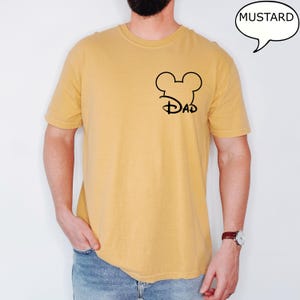 May include: Mustard-colored t-shirt with a black Mickey Mouse head and "Dad" design. The shirt is short-sleeved and has a crew neck. A speech bubble with the word "Mustard" is in the upper right corner.
