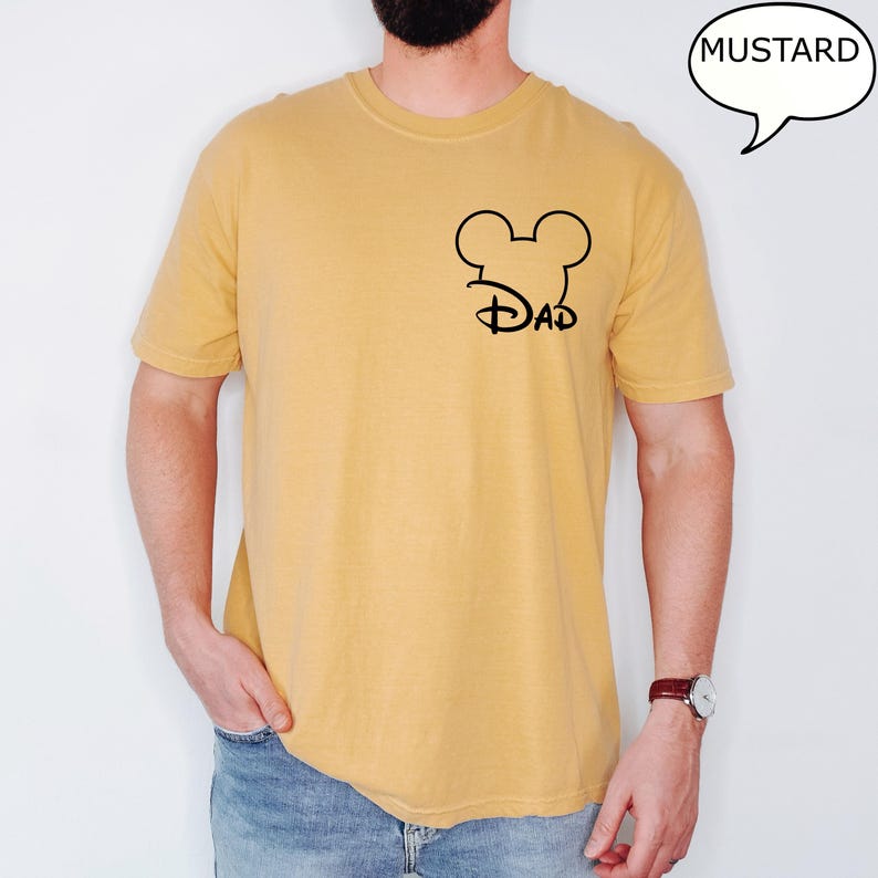 May include: Mustard-colored t-shirt with a black Mickey Mouse head and "Dad" design. The shirt is short-sleeved and has a crew neck. A speech bubble with the word "Mustard" is in the upper right corner.