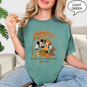 May include: Light green t-shirt featuring a graphic of Mickey and Minnie Mouse embracing under a tree with autumn leaves. The text "It's the most wonderful time of the year" is printed below the image. The shirt is a solid color.
