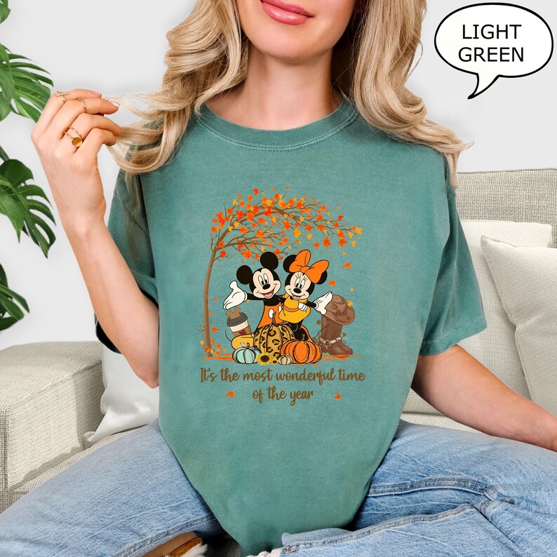 May include: Light green t-shirt featuring a graphic of Mickey and Minnie Mouse embracing under a tree with autumn leaves. The text "It's the most wonderful time of the year" is printed below the image. The shirt is a solid color.