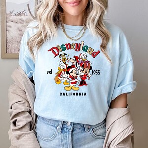 May include: Light blue t-shirt with a graphic of Mickey Mouse and friends in front of a Disneyland logo. The logo says "Disneyland est. 1955 California".
