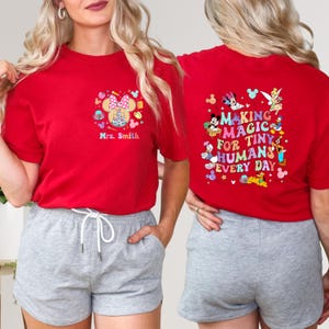 May include: Red t-shirt with a colourful graphic on the front featuring a castle, Minnie Mouse ears, and the text "Mrs. Smith." The back of the shirt has the text "MAKING MAGIC FOR TINY HUMANS EVERY DAY" with cartoon characters.