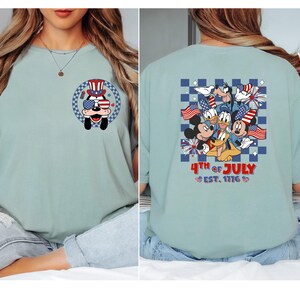 Pode incluir: Camiseta azul claro com um cachorro de desenho animado usando &oacute;culos de sol e chap&eacute;u patri&oacute;ticos. A parte de tr&aacute;s da camisa mostra personagens de desenho animado com bandeiras americanas e fogos de artif&iacute;cio, com o texto "4th of July Est. 1776."