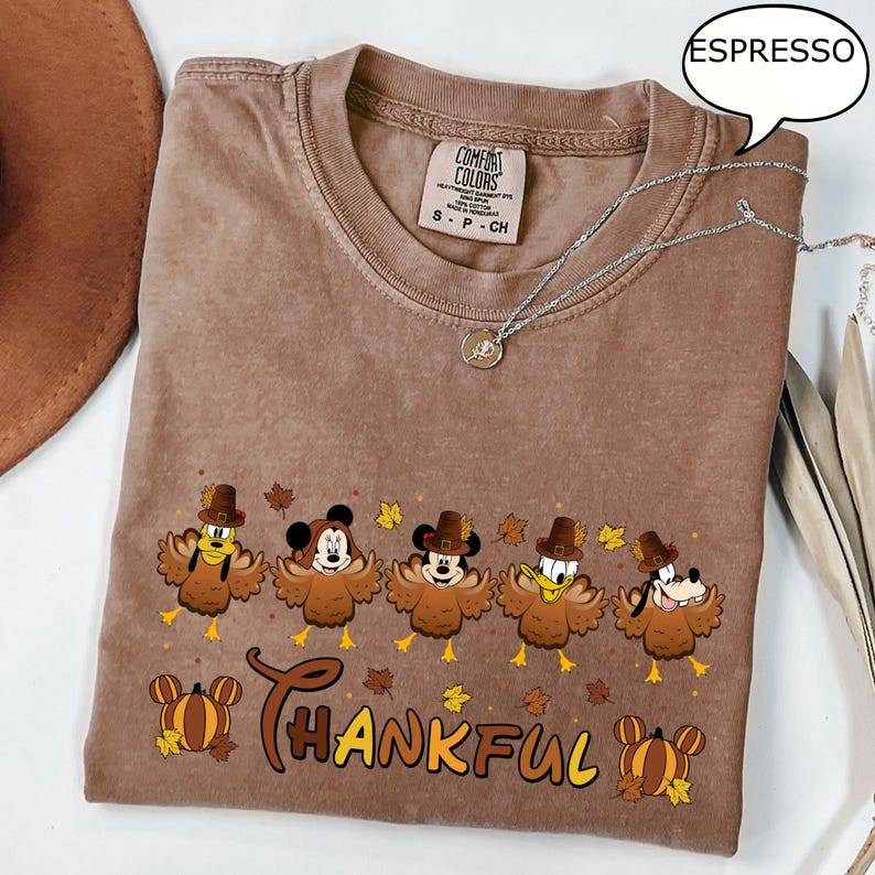 May include: Espresso-colored t-shirt with a graphic of cartoon turkeys wearing pilgrim hats. The word "THANKFUL" is printed below the turkeys. A silver necklace with a pendant is draped across the shirt.