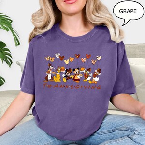 May include: A purple t-shirt featuring a Thanksgiving design with cartoon characters and Mickey Mouse-shaped balloons. The word "THANKSGIVING" is printed in brown across the bottom. The shirt is a comfortable, casual style.