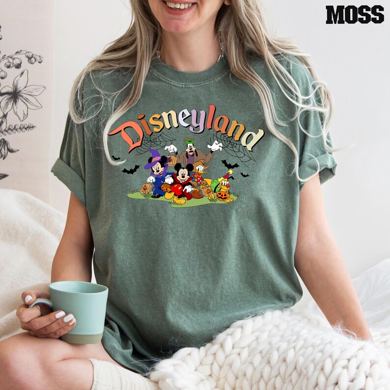 Puede incluir: Una camiseta verde con el texto "Disneyland" en colores arco&iacute;ris. La camiseta presenta un gr&aacute;fico de Mickey Mouse, Minnie Mouse, Goofy, Donald Duck y Pluto vestidos con disfraces de Halloween.