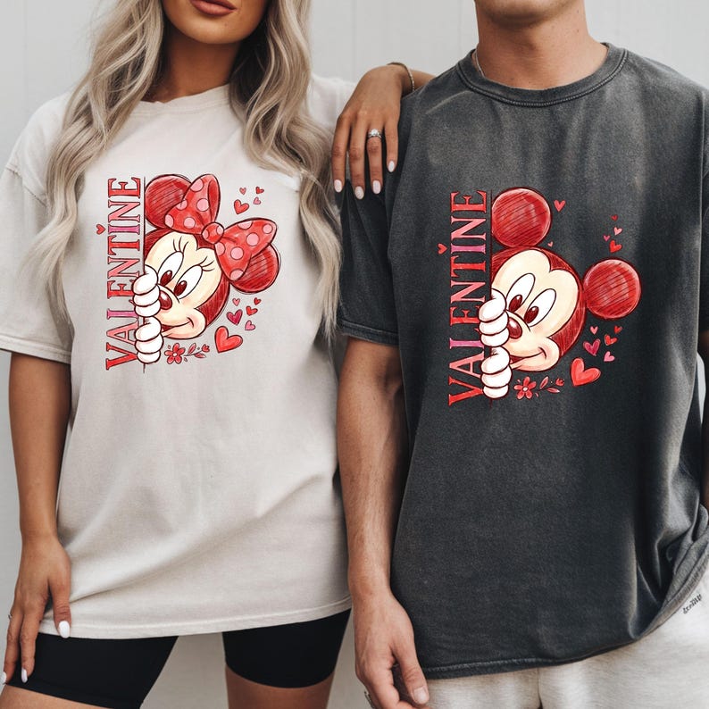 Puede incluir: Dos camisetas holgadas, una crema y otra carb&oacute;n, con un dise&ntilde;o de San Valent&iacute;n. El dise&ntilde;o incluye un rat&oacute;n de dibujos animados con un lazo rojo y corazones, con la palabra "VALENTINE" impresa verticalmente en rojo.