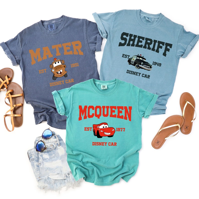 Puede incluir: Tres camisetas con tem&aacute;tica de Cars. La camiseta azul dice "MATER EST 1951 DISNEY CAR" con una imagen de un cami&oacute;n de remolque rojo. La camiseta azul claro dice "SHERIFF EST 1949 DISNEY CAR" con una imagen de un coche de polic&iacute;a azul. La camiseta turquesa dice "MCQUEEN EST 1977 DISNEY CAR" con una imagen de un coche de carreras rojo.