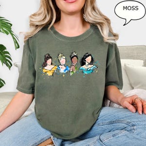 May include: A moss green t-shirt featuring a watercolor illustration of four princesses. The princesses are depicted with various skin tones and hairstyles, wearing gowns in yellow, blue, and teal. The word "MOSS" is in a speech bubble.