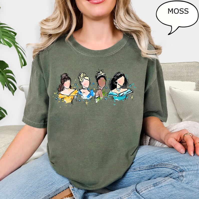 May include: A moss green t-shirt featuring a watercolor illustration of four princesses. The princesses are depicted with various skin tones and hairstyles, wearing gowns in yellow, blue, and teal. The word "MOSS" is in a speech bubble.